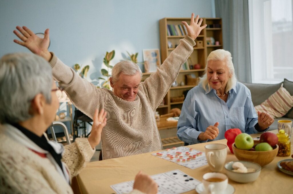 Group of elderly friends enjoying each other's company while playing a board game around dining table, with cheerful expressions and raised hands in celebration